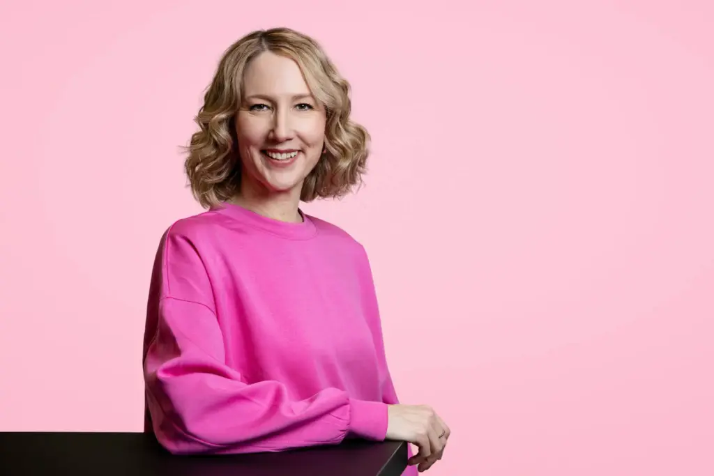 corine brötz in pinkfarbenem outfit vor pinkem hintergrund, freundlich lächelnd, blickt in die kamera, arm auf dem tisch – professionelles business portrait
