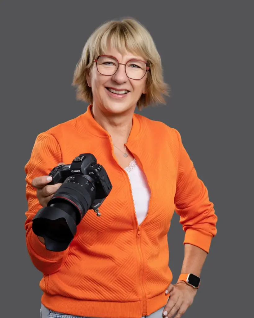 Karina Schuh mit Kamera und orangefarbener Jacke mit grauem Hintergrund im Studio in Polch bei Koblenz