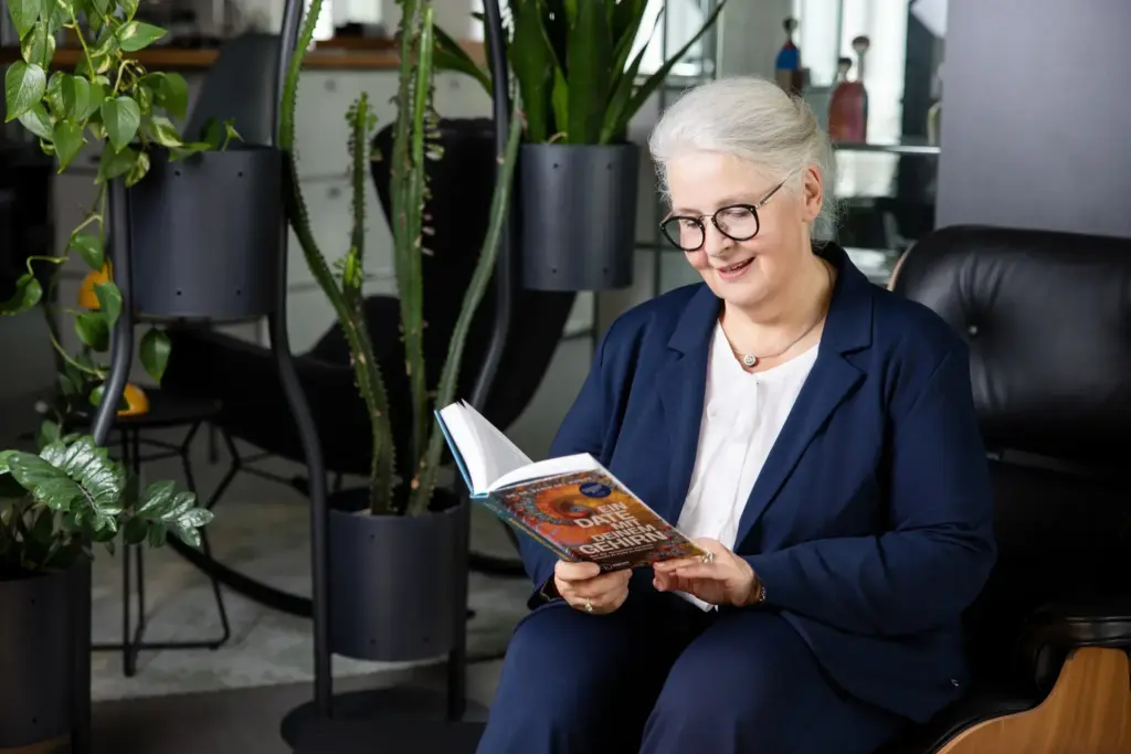 Dagmar Gösling sitzt zwischen grünen Pflanzen bei designfunktion Koblenz und liest in einem Fachbuch