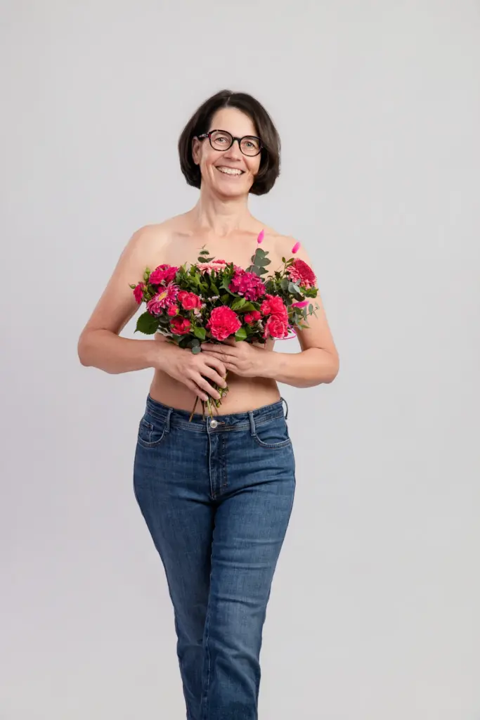 Fröhliche Frau mit Brille in Jeans hält pinke Blumen vor der Brust, Portrait für die Pinktober-Plakatkampagne Koblenz 2026