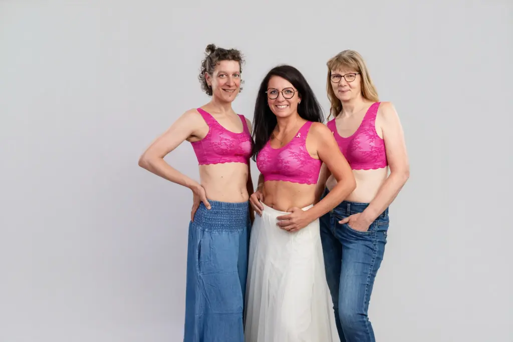 Drei Frauen in pinken BHs, zwei in Jeans, eine im weißen Tüllrock, stehen fröhlich zusammen, Pinktober Koblenz 2026