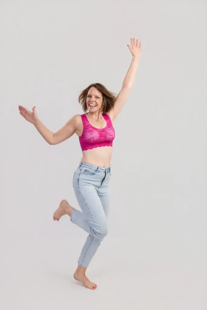 Frau im pinken BH und hellen Jeans springt lachend mit ausgebreiteten Armen, Pinktober Koblenz 2026
