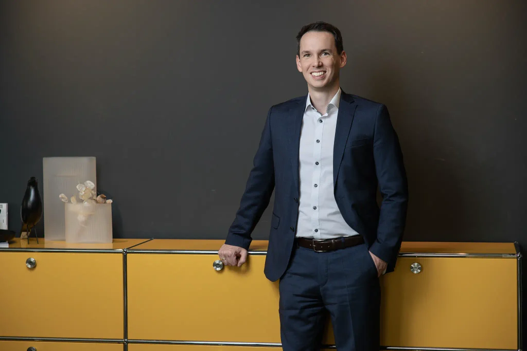 Businessfotos Steuerberater: Tobias Junges im dunklen Anzug vor dunkler Wand und gelbem Sideboard bei designfunktion