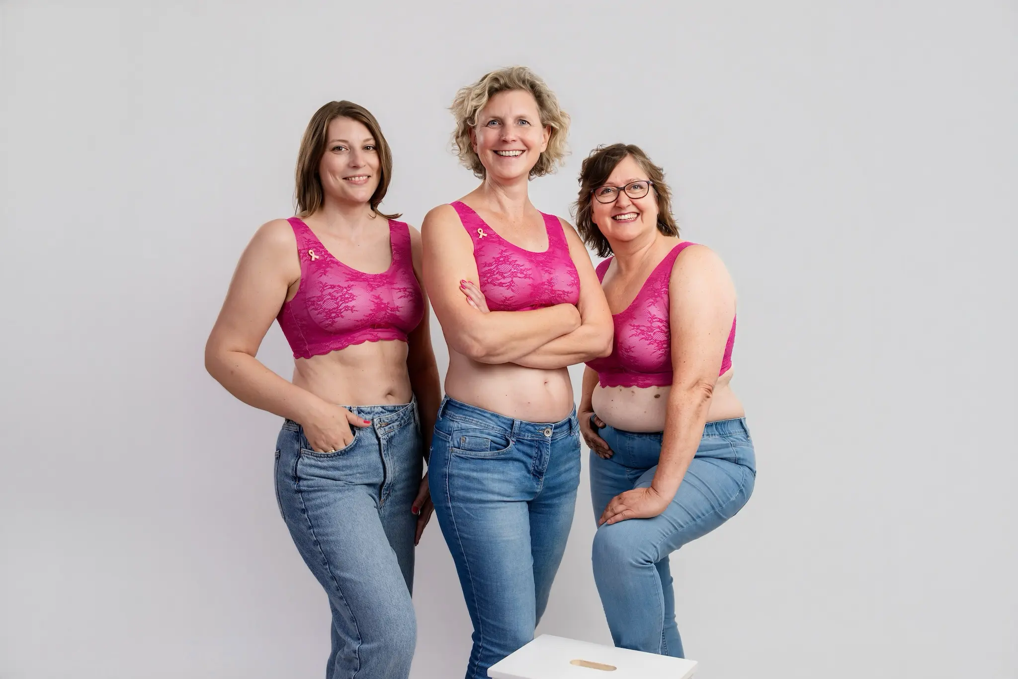 Drei lächelnde Frauen in pinkfarbenen Spitzen-Bralettes und Jeans stehen eng beieinander vor grauem Hintergrund, Fotoshooting für Frauen mit Krebs in Koblenz